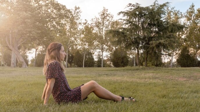 A young woman with long hair sits on a grassy lawn, gazing into the distance with a blurred background.