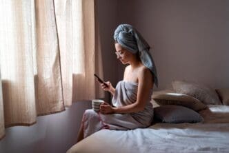A young woman sits on a bed, looking at her phone.