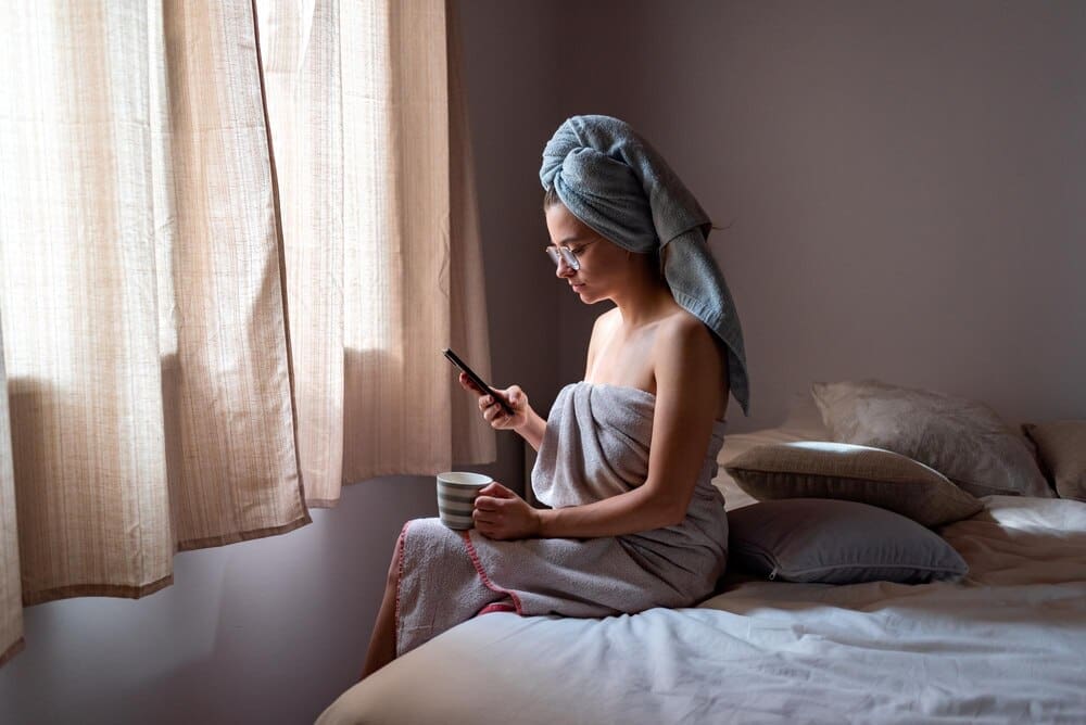 A young woman sits on a bed, looking at her phone.
