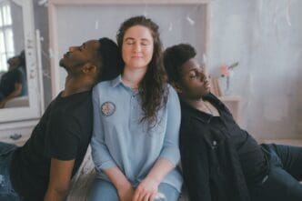 A woman who appears to be White sits with her eyes closed, flanked by two men who appear to be Black, also with closed eyes, leaning against her. All three appear calm or introspective in an indoor setting.