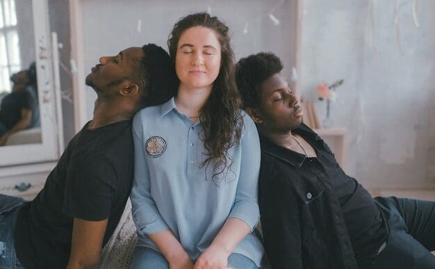 A woman who appears to be White sits with her eyes closed, flanked by two men who appear to be Black, also with closed eyes, leaning against her. All three appear calm or introspective in an indoor setting.