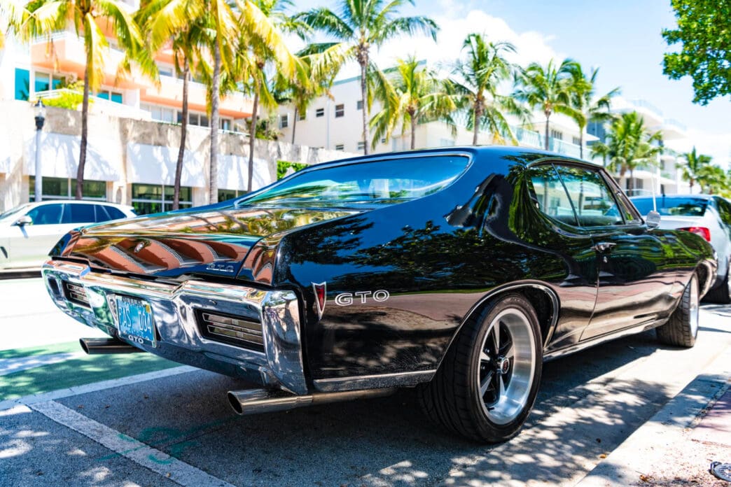 A classic black 1968 Pontiac GTO with a sleek design and chrome bumper is parked on a street in Miami Beach, with palm trees and a hotel in the background.