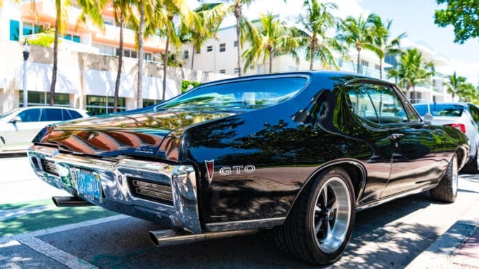A classic black 1968 Pontiac GTO with a sleek design and chrome bumper is parked on a street in Miami Beach, with palm trees and a hotel in the background.