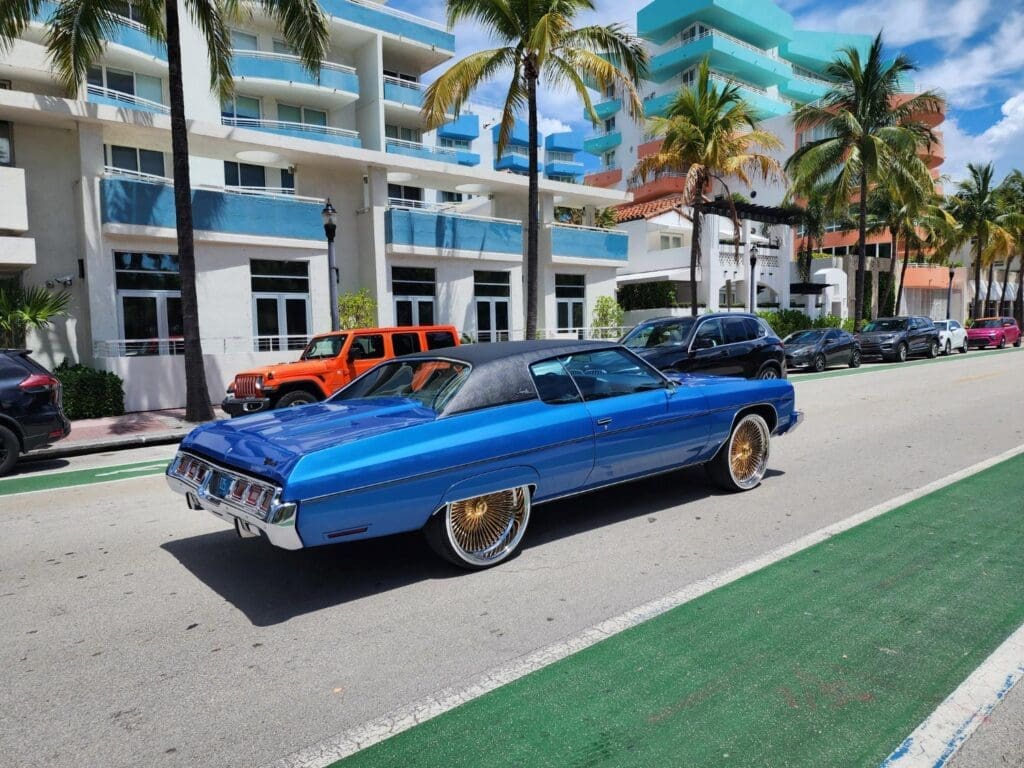 A classic blue 1973 Chevrolet Impala Coupe with gold spoked rims is driving on a street in Miami Beach, with art deco buildings and palm trees in the background.