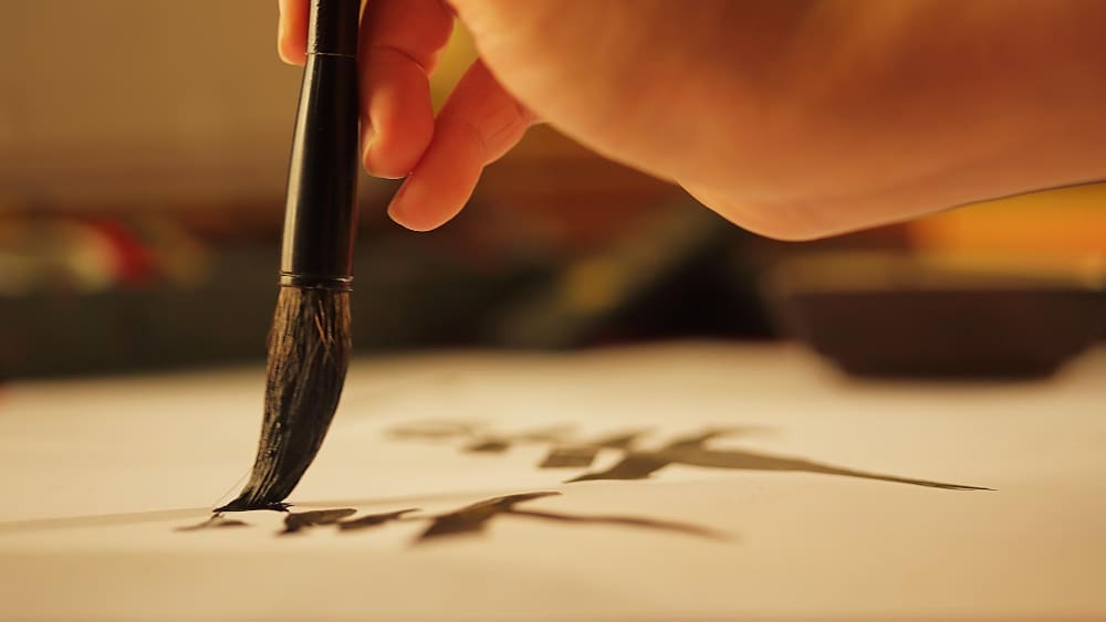 A close-up of a hand holding a traditional calligraphy brush, writing an ink character on white paper.