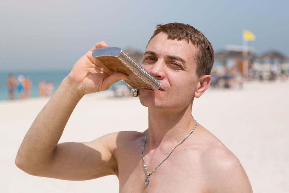 A man in his thirties or forties drinks from a metal flask while sitting on a city beach.