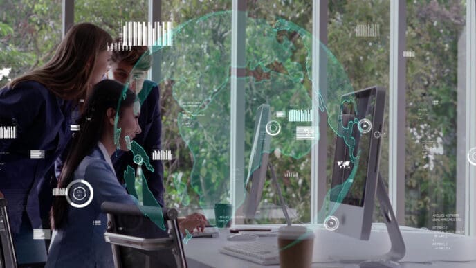 Three professionals, one seated and two standing, look at a computer screen in an office with large windows, overlaid with holographic digital graphics of a world map and data charts, symbolizing the integration of AI into the workplace.