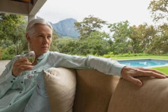 A serious-looking senior woman with grey hair holds a glass of white wine while sitting on a porch, with a swimming pool and mountains in the lush green background.