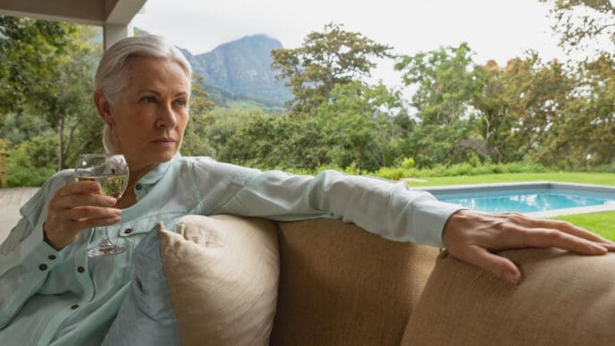 A serious-looking senior woman with grey hair holds a glass of white wine while sitting on a porch, with a swimming pool and mountains in the lush green background.