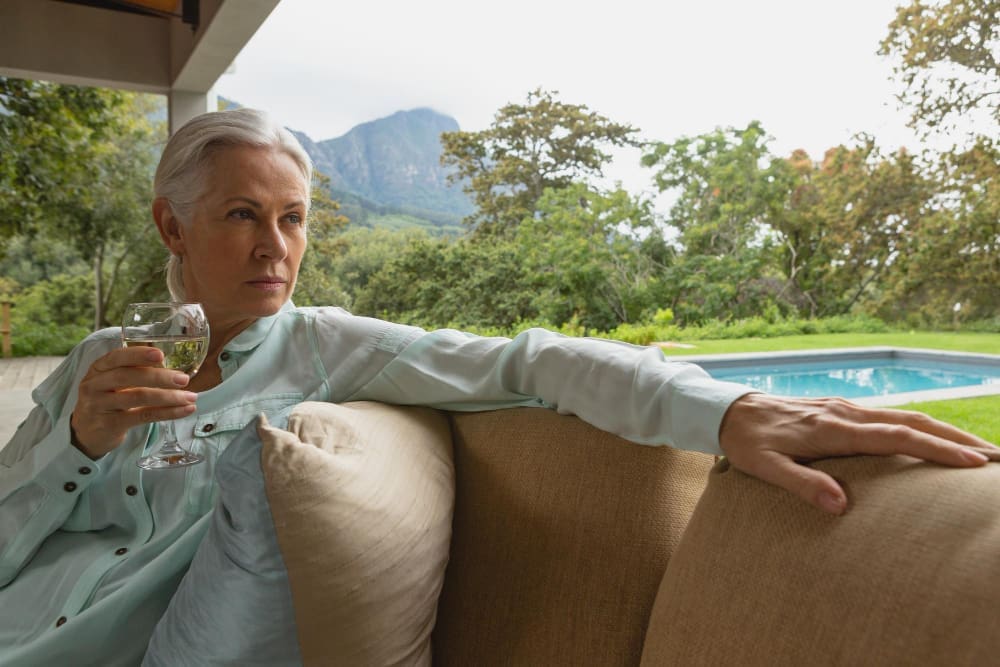 A serious-looking senior woman with grey hair holds a glass of white wine while sitting on a porch, with a swimming pool and mountains in the lush green background.