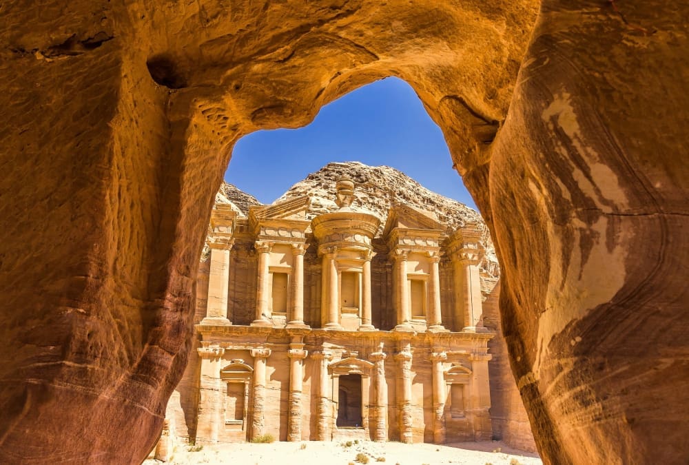 The Monastery (Ad Deir) in Petra, Jordan, is seen from a cave opening, with the ancient building carved into the rock face.