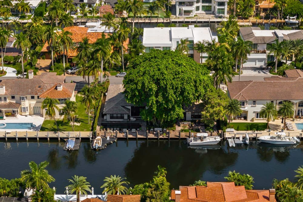 An aerial view of a luxurious residential neighborhood in Coral Gables, Florida, with a waterway lined with docks and boats, large houses, and lush palm trees, under a clear sky.