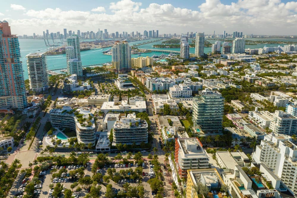 An expansive aerial view of Miami Beach, showcasing a dense urban landscape of mid-rise and high-rise buildings, with the distinct downtown Miami skyline and causeways visible across the turquoise bay in the distance under a clear blue sky.