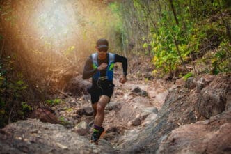 A male trail runner in a blue vest and a cap is running on a rocky path through a dense, green forest, with bright sunlight shining through the trees, in a race known as Akha Trail (THA).