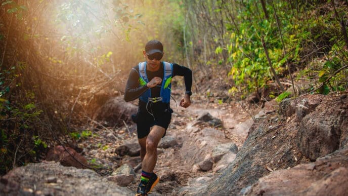 A male trail runner in a blue vest and a cap is running on a rocky path through a dense, green forest, with bright sunlight shining through the trees, in a race known as Akha Trail (THA).