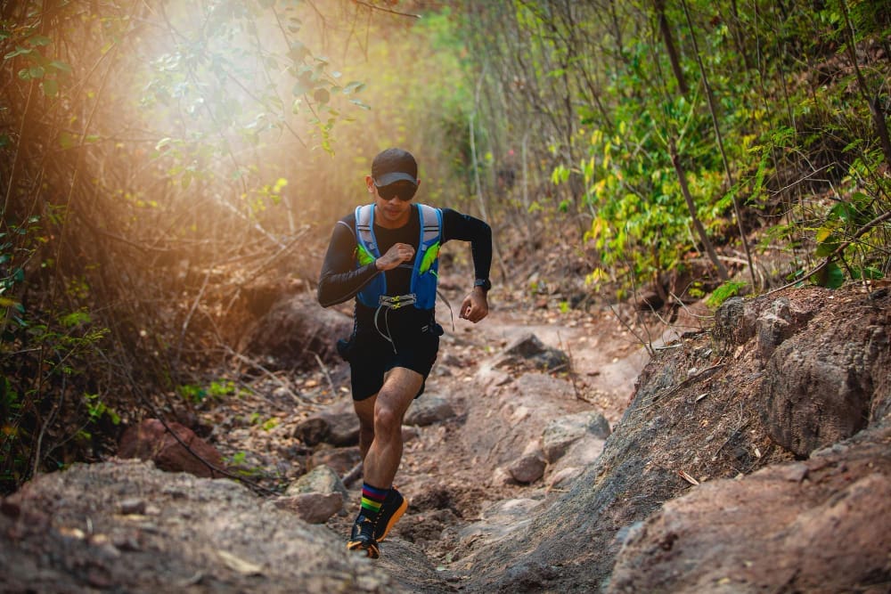 A male trail runner in a blue vest and a cap is running on a rocky path through a dense, green forest, with bright sunlight shining through the trees, in a race known as Akha Trail (THA).