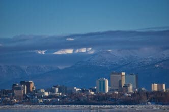The skyline of Anchorage, Alaska, is seen with a frozen bay in the foreground and a snow-capped mountain range in the background.