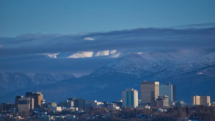 The skyline of Anchorage, Alaska, is seen with a frozen bay in the foreground and a snow-capped mountain range in the background.
