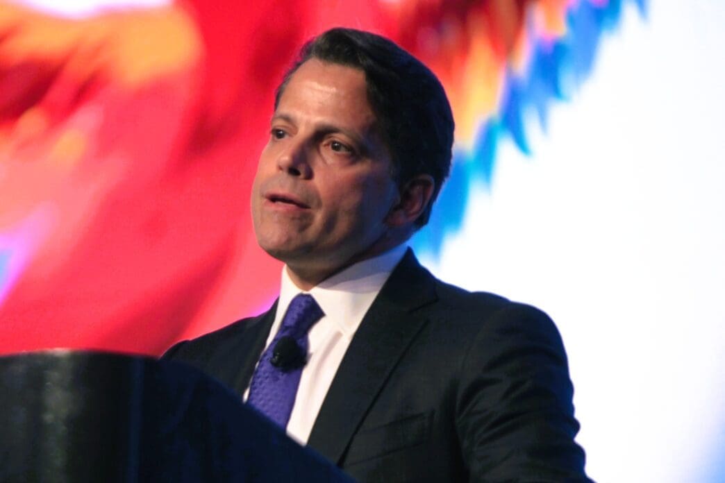 A photograph of Anthony Scaramucci, a man in a dark suit, speaking at a podium in front of a colorful, blurred background.