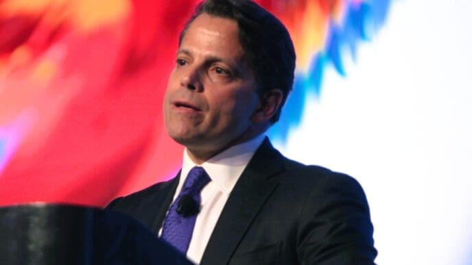 A photograph of Anthony Scaramucci, a man in a dark suit, speaking at a podium in front of a colorful, blurred background.