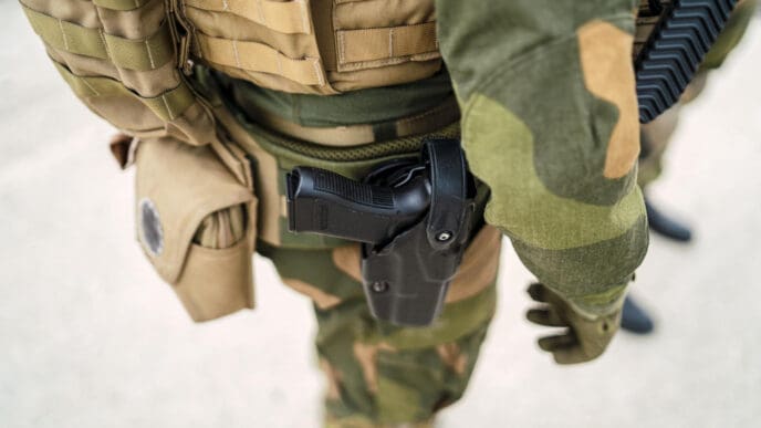 A close-up, high-angle view of an armed National Guard soldier in a camouflage uniform, with a handgun holstered on their right hip.