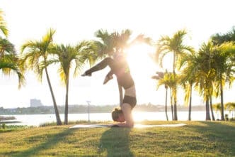 A woman performs a yoga forearm stand pose on a mat on a grassy lawn, with the sun setting behind her and palm trees in the background.
