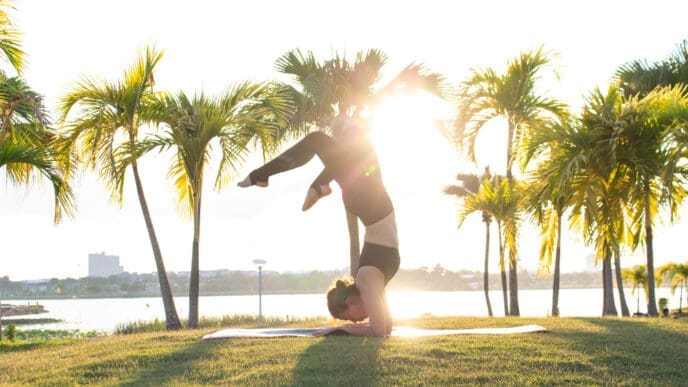A woman performs a yoga forearm stand pose on a mat on a grassy lawn, with the sun setting behind her and palm trees in the background.