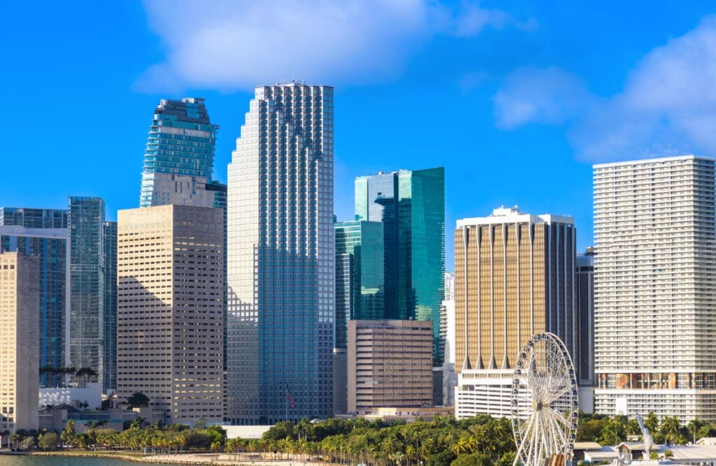 A panoramic view of the Miami skyline with a tall Ferris wheel and Biscayne Bay in the foreground.