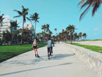 A man on a bicycle and a woman rollerblading are moving down a wide concrete path, surrounded by palm trees, green grass, and a beach, with buildings in the background under a clear blue sky.