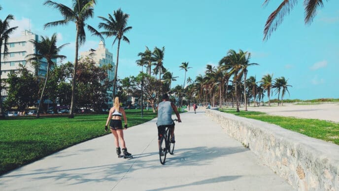 A man on a bicycle and a woman rollerblading are moving down a wide concrete path, surrounded by palm trees, green grass, and a beach, with buildings in the background under a clear blue sky.