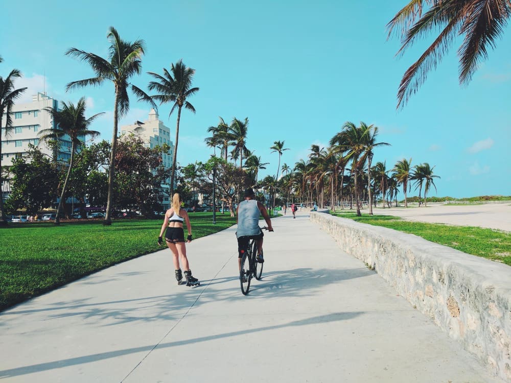 A man on a bicycle and a woman rollerblading are moving down a wide concrete path, surrounded by palm trees, green grass, and a beach, with buildings in the background under a clear blue sky.