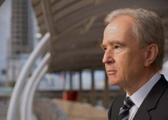 A close-up, side-profile portrait of a serious-looking Bernard Arnault in a business suit against a blurred urban background.