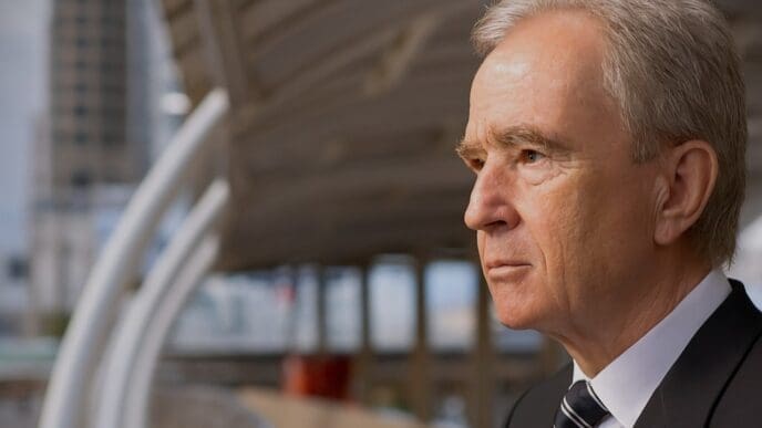 A close-up, side-profile portrait of a serious-looking Bernard Arnault in a business suit against a blurred urban background.