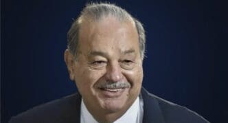 A close-up portrait of billionaire Carlos Slim, smiling and wearing a suit against a blue background.