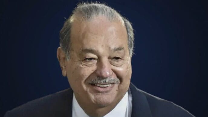 A close-up portrait of billionaire Carlos Slim, smiling and wearing a suit against a blue background.