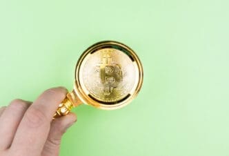 A person's hand holds a golden magnifying glass over a golden Bitcoin coin, all against a plain green background.