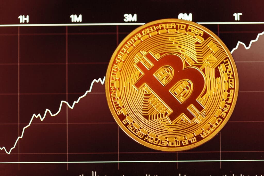 A close-up of a golden Bitcoin coin overlaid on a financial chart with a white line showing an upward price trend.