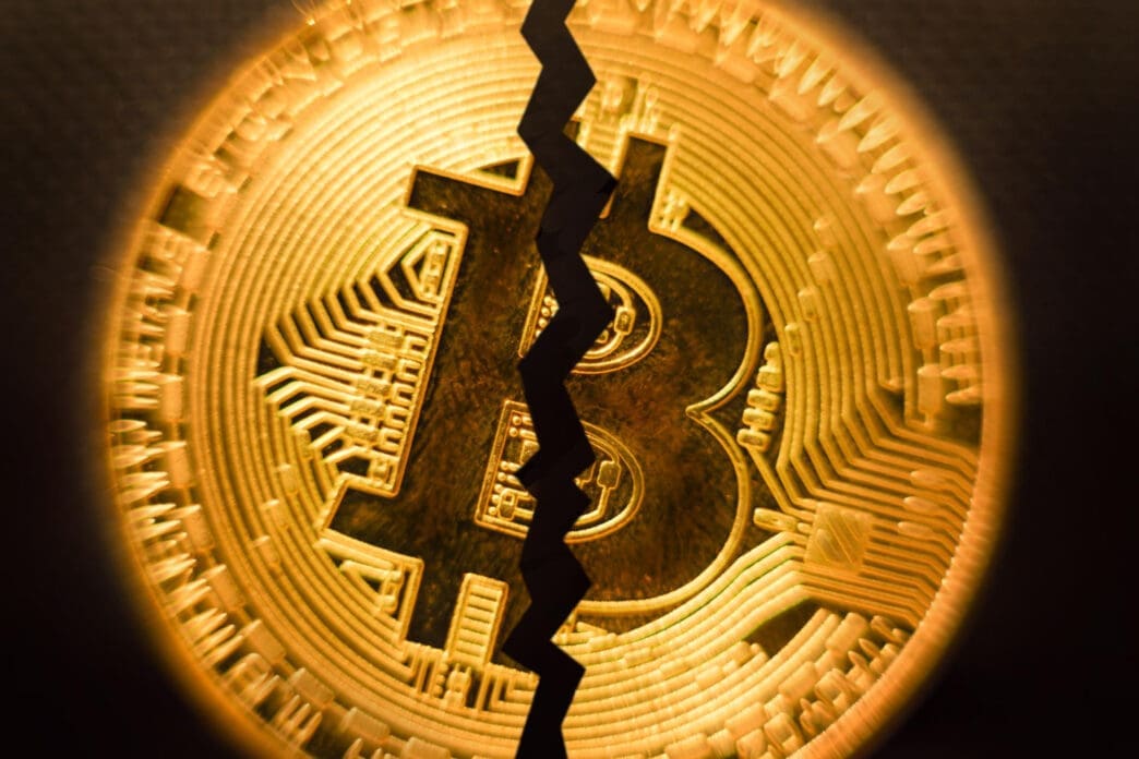 A close-up photograph of a golden Bitcoin coin with a prominent, jagged black crack running vertically through its center.