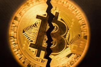 A close-up photograph of a golden Bitcoin coin with a prominent, jagged black crack running vertically through its center.