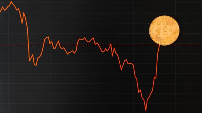 A financial chart shows a sharp downward trend, with a Bitcoin coin placed at the end of the line on the right.