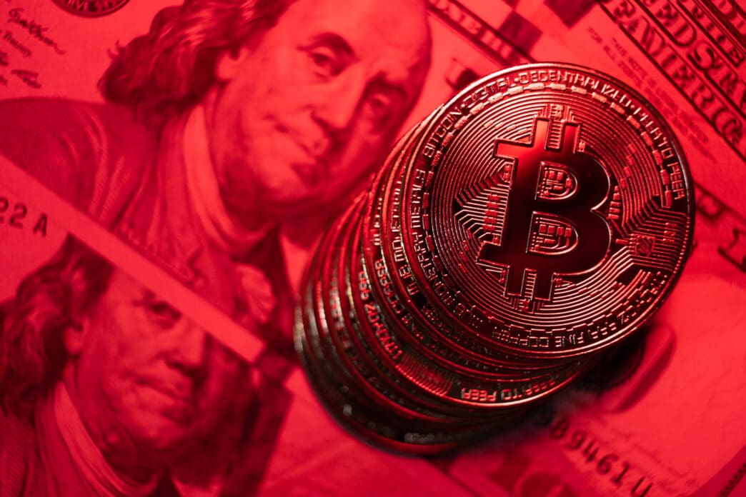 A close-up photograph of a stack of golden Bitcoin coins illuminated by a dramatic red light, with a blurry US hundred-dollar bill visible in the background.
