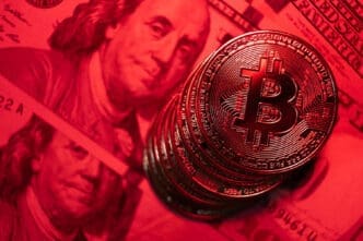 A close-up photograph of a stack of golden Bitcoin coins illuminated by a dramatic red light, with a blurry US hundred-dollar bill visible in the background.