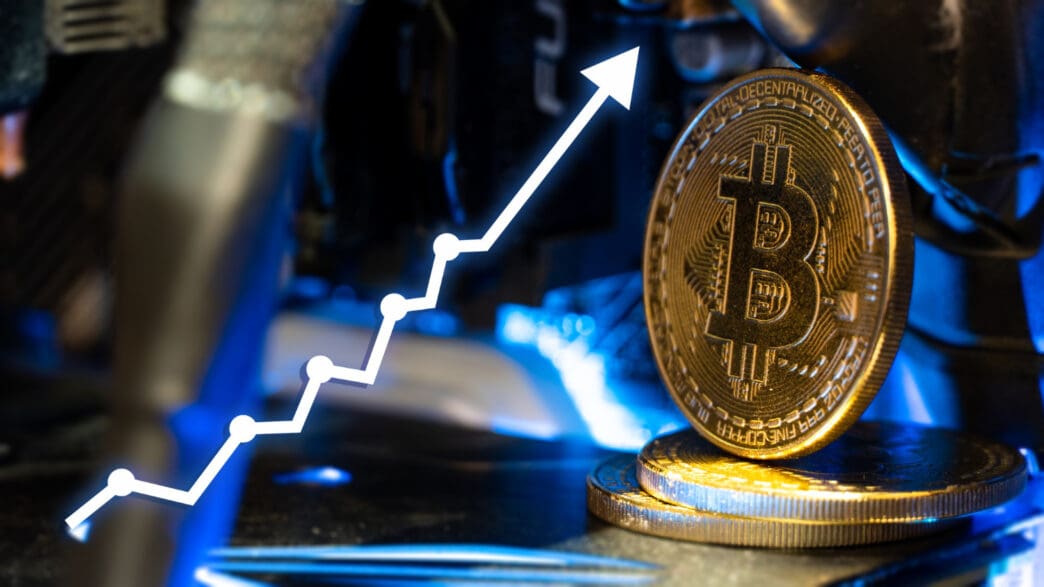 A golden Bitcoin coin stands on its edge on computer hardware, with a white, digitally overlaid line graph showing an upward trend.