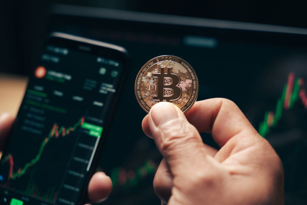 A hand holds a Bitcoin coin over a smartphone and a laptop displaying a financial chart