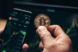 A hand holds a Bitcoin coin over a smartphone and a laptop displaying a financial chart