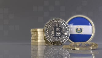 A stack of gold coins and a Bitcoin coin stand next to a round emblem of the flag of El Salvador.