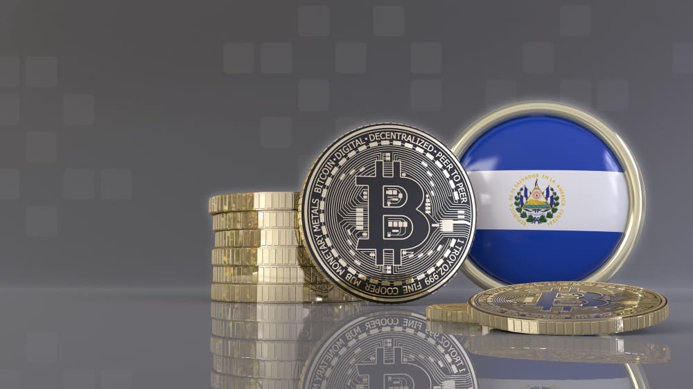 A stack of gold coins and a Bitcoin coin stand next to a round emblem of the flag of El Salvador.