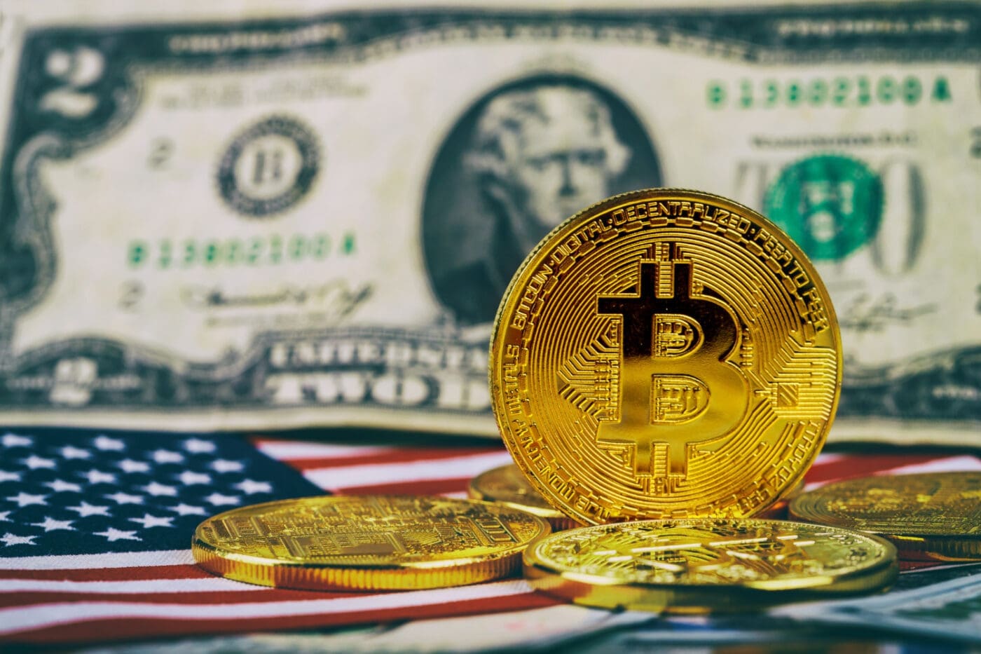 A golden Bitcoin coin stands upright on a surface, with other Bitcoin coins, a US two-dollar bill, and a portion of the American flag visible in the background.