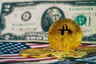 A golden Bitcoin coin stands upright on a surface, with other Bitcoin coins, a US two-dollar bill, and a portion of the American flag visible in the background.