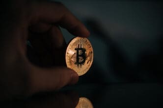 A close-up photograph of a hand holding a single, shiny golden Bitcoin coin, which is reflected on a dark surface below.
